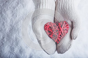 Female hands in white knitted mittens with entwined vintage romantic red heart on snow background. Love and St. Valentine concept