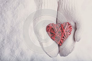 Female hands in white knitted mittens with a entwined vintage romantic red heart on a snow. Love and St. Valentine concept.