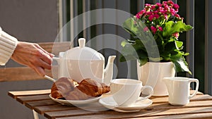 Female hands in a white sweater pour tea from a white teapot into a white ceramic cup outside, a table served with