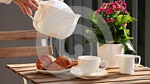Female hands in a white sweater pour tea from a white teapot into a white ceramic cup outside, a table served with