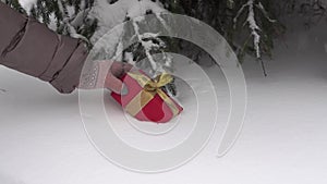 Female hands in winter gloves put a Christmas gift box under the tree in a snowy winter forest. Fluffy snow is falling.