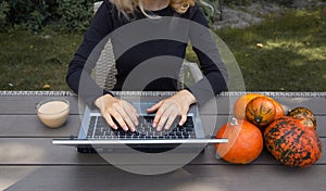 Female hands are working on a laptop. Next to keyboard is cup of cappuccino and small orange pumpkins