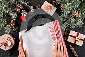 Female hands wrapping a Christmas gift on holiday wrapping on black background with fir branches, red decorations