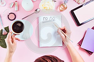 Female hands write To do list on the pink background with cosmetics, coffee cup, notebooks, tablet with blank screen. Beauty blog