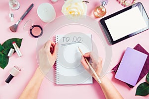 Female hands write To do list on the pink background with cosmetics, coffee cup, notebooks, tablet with blank screen. Beauty blog