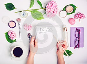 Female hands write To do list on the white working place with female accessories, cup of coffee, notebook, glasses, and wisteria f