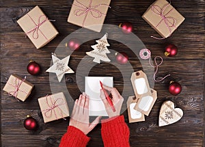 Female hands writing christmas gift list on paper on wooden background with gifts and labels