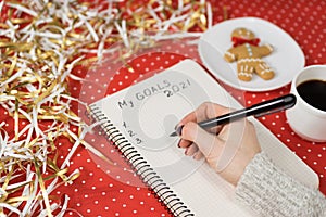Female hands writing My Goals 2021 in a notebook. Gingerbread man, coffee, red background and tinsel. New Years concept