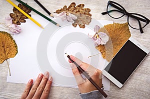 Female hands writing on white blank paper on wooden table with pencils, mobile phone, glasses and autumn leaves.