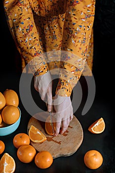 Female hands in a yellow dress cuts out an orange on a cutting board on a dark background.