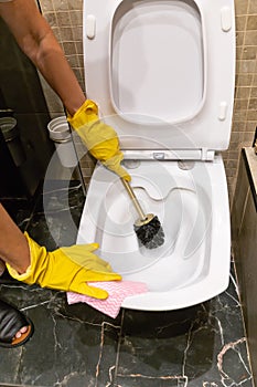 Female hands in yellow gloves cleaning the toilet bowl with brush and rag in a bathroom, vertical
