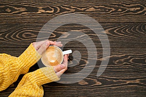 Female hands in yellow sweater holding a cup of coffee on the wooden background. Top view of hot cappuccino on the table.