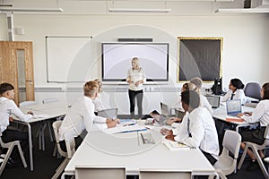 Female High School Teacher Standing Next To Interactive Whiteboard And Teaching Lesson To Pupils Wearing Uniform photo