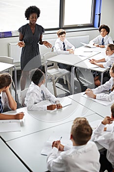 Female High School Tutor Helping Students Wearing Uniform Seated Around Tables In Lesson