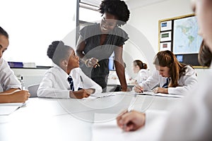 Female High School Tutor Helping Students Wearing Uniform Seated Around Tables In Lesson