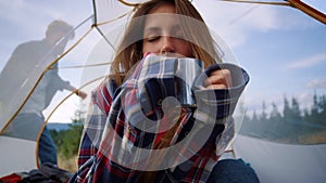 Female hiker drinking tea from mug in tent. Girl and guy sitting in tourist tent