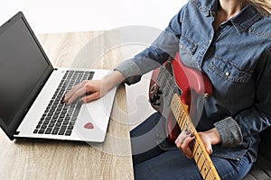 Casually dressed young woman with guitar playing songs in the room at home. Laptop on table. Online guitar lessons concept. Male g