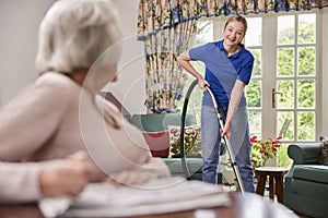 Female Home Help Cleaning House With Vacuum Cleaner And Talking To Senior Woman