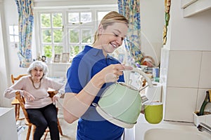 Female Home Help Making Cup Of Tea In Kitchen Whilst Chatting With Senior Woman