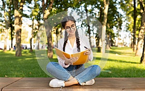 Female indian student girl writing notes to notebook while sitting on bench in university campus or park