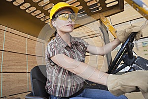 Female industrial worker driving forklift truck with stacked wooden planks in background