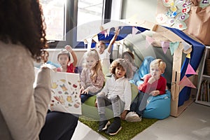 Female infant school teacher sitting on a chair showing a book to a group of children sitting on bean bags in a comfortable corner