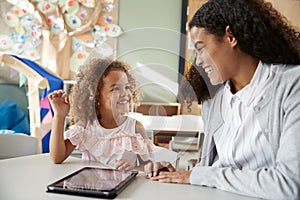Female infant school teacher working one on one in a classroom using a tablet computer with a young mixed race schoolgirl, smiling