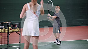 Female instructor teaching little boy playing tennis. Woman in white sport outfit standing in the court throwing yellow