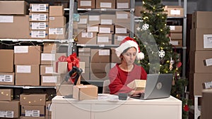 Female inventory manager wearing Santa hat using laptop computer to check order number on a parcel package, preparing