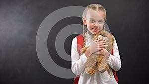 Female kid with rucksack hugging teddy bear smiling on camera against blackboard