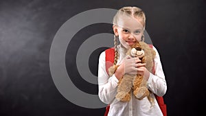 Female kid with rucksack hugging teddy bear smiling on camera against blackboard