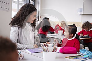 Female kindergarten teacher sitting at table in a classroom talking to a young Chinese schoolgirl, close up