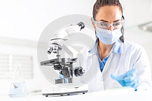 Female lab technician in protective glasses, gloves and face mask sits next to a microscope and conical flask, looking aside on