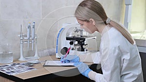 a female laboratory assistant studies microorganisms by looking through a microscope and writes the readings in a log.