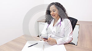 Female latin female doctor sitting looking at her phone in her office with stethoscope on her neck typing on her smartphone