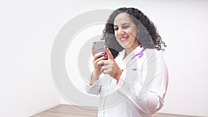 Female latin female doctor smiling standing looking at her phone in her office with stethoscope on her neck typing on her phone