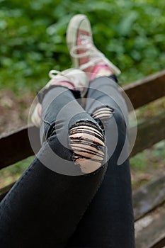 Female legs in black ripped jeans and pink canvas sneakers lifted on the bench