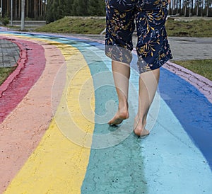 Female legs close up on barefoot on wet asphalt drawn rainbow walking path after rain among puddles, copy space, love, freedom and