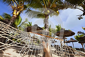 Female legs on hammock on tropical beach with palm leaf thatch roofing umbrellas and palm trees in the background