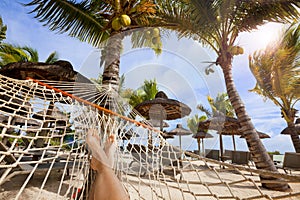 Female legs on hammock on tropical beach with palm leaf thatch roofing umbrellas and palm trees in the background