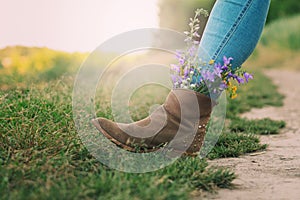 Female legs in jeans high boots with inserted flowers on the road in the field.