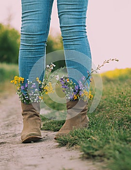 Female legs in jeans high boots with inserted flowers on the road in the field