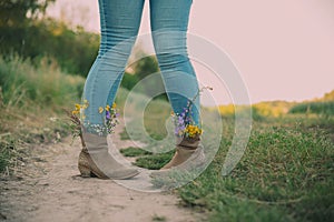 Female legs in jeans high boots with inserted flowers on the road in the field