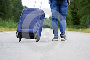 Female legs in jeans and sneakers with a travel bag on wheels walks away on the road