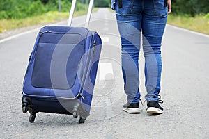 Female legs in jeans and sneakers with a travel bag on wheels walks away on the road