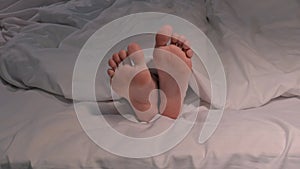 female legs joyfully movie and dance with toes while lying in bed on a sheet under blanket. Happy woman girl with bare