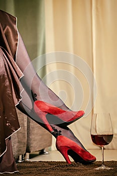 female legs in red suede high-heeled shoes and a glass of wine on the floor in the room