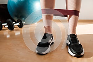 Female legs in sports leggings and sneakers doing exercises with fitness elastic bands at home during lockdown. Home