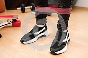 Female legs in sports leggings and sneakers doing exercises with fitness elastic bands at home during lockdown. Home