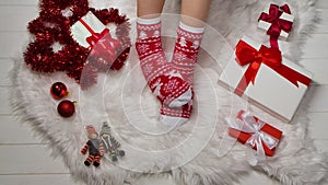 Female legs in warm socks with red white ornaments on a background of fur skin, tinsel and boxes with gifts relaxing at